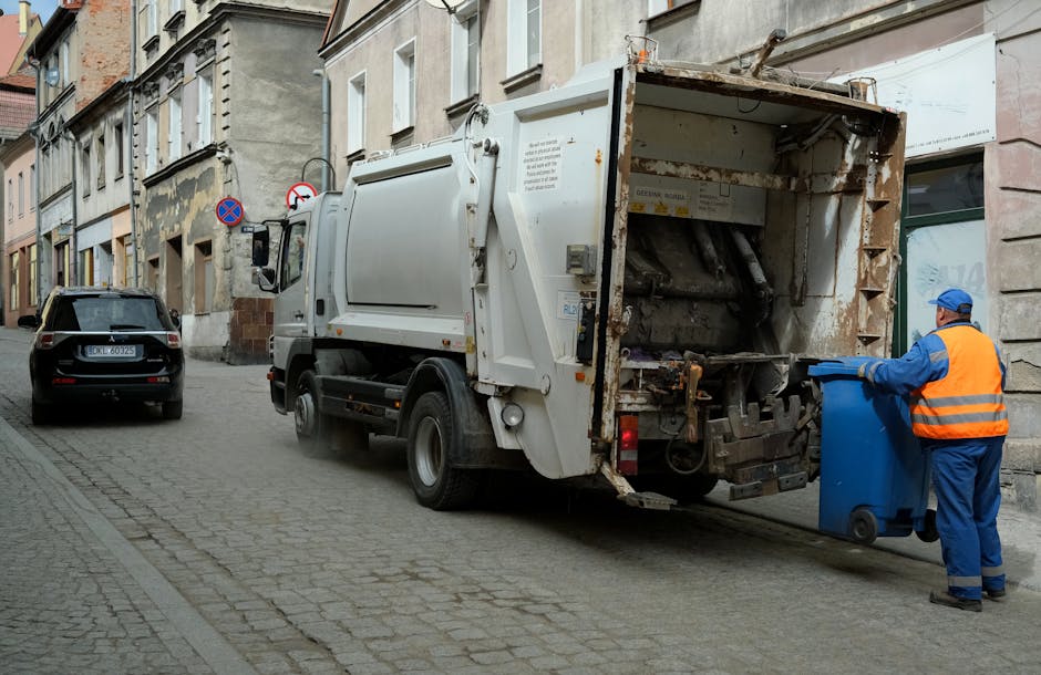 A large collection of mixed domestic rubbish and waste materials is piled and overflowing from multiple wheelie bins located on a paved sidewalk area in front of a retail shopping centre or commercial