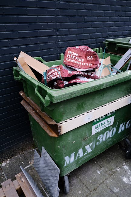 A person is placing a crumpled brown paper bag into a white, open rubbish bin with a black interior, situated on a wooden surface. The person’s hand is visible, gripping the bag by its top edge, and