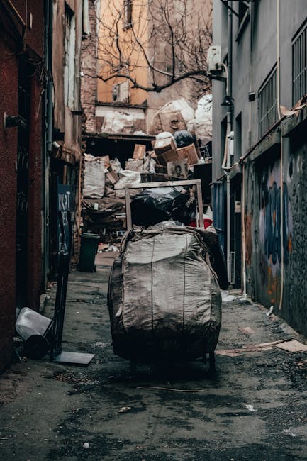 A pile of discarded cardboard boxes, some flattened and others partially assembled or torn, is shown stacked against a concrete block wall outdoors. The boxes vary in size and are made of corrugated b
